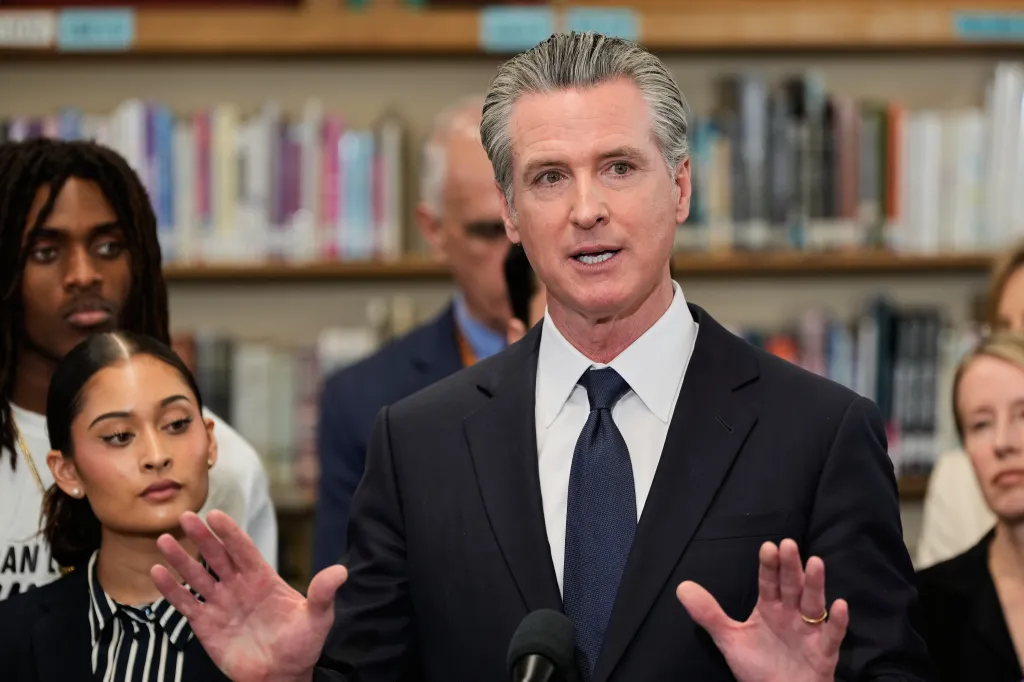 Governor Gavin Newsom speaking at a press conference, gesturing with his hands.