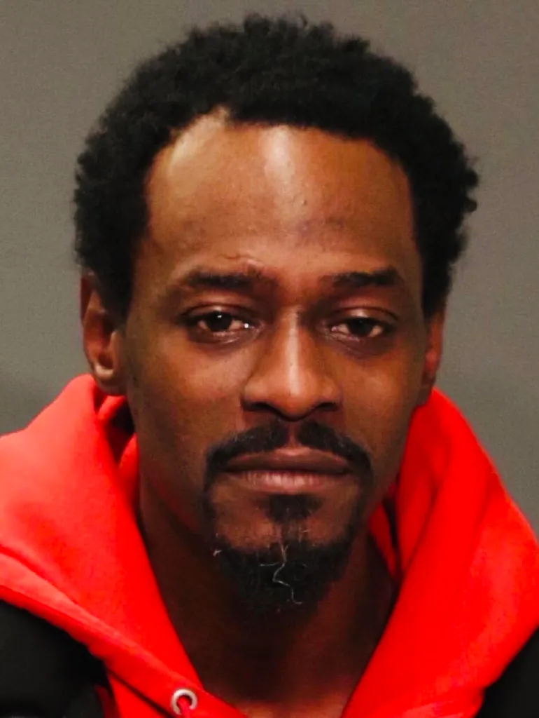 Mugshot of a man with dark skin, a close-cut afro, a dark goatee, and wearing a red hoodie.