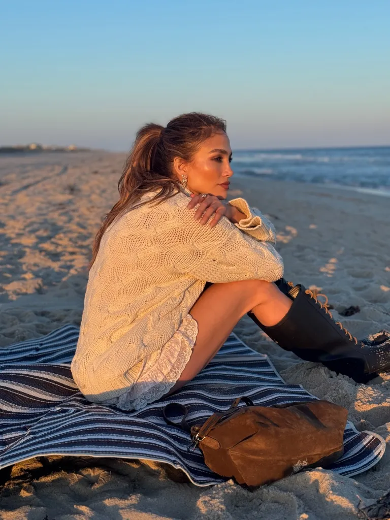 Jennifer Lopez sits on a striped blanket on the beach at sunset, looking out at the ocean.