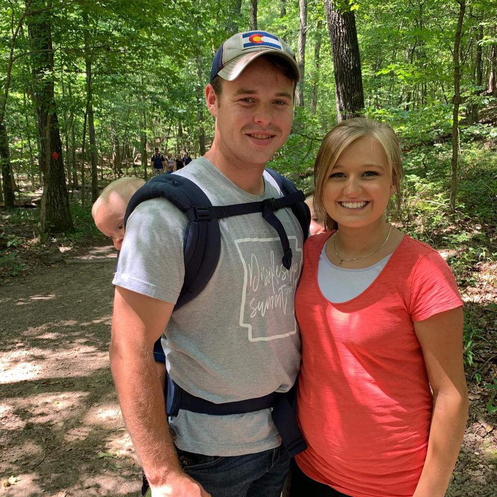 Joseph and Kendra Duggar smiling while Joseph carries a child in a baby carrier on a hike.