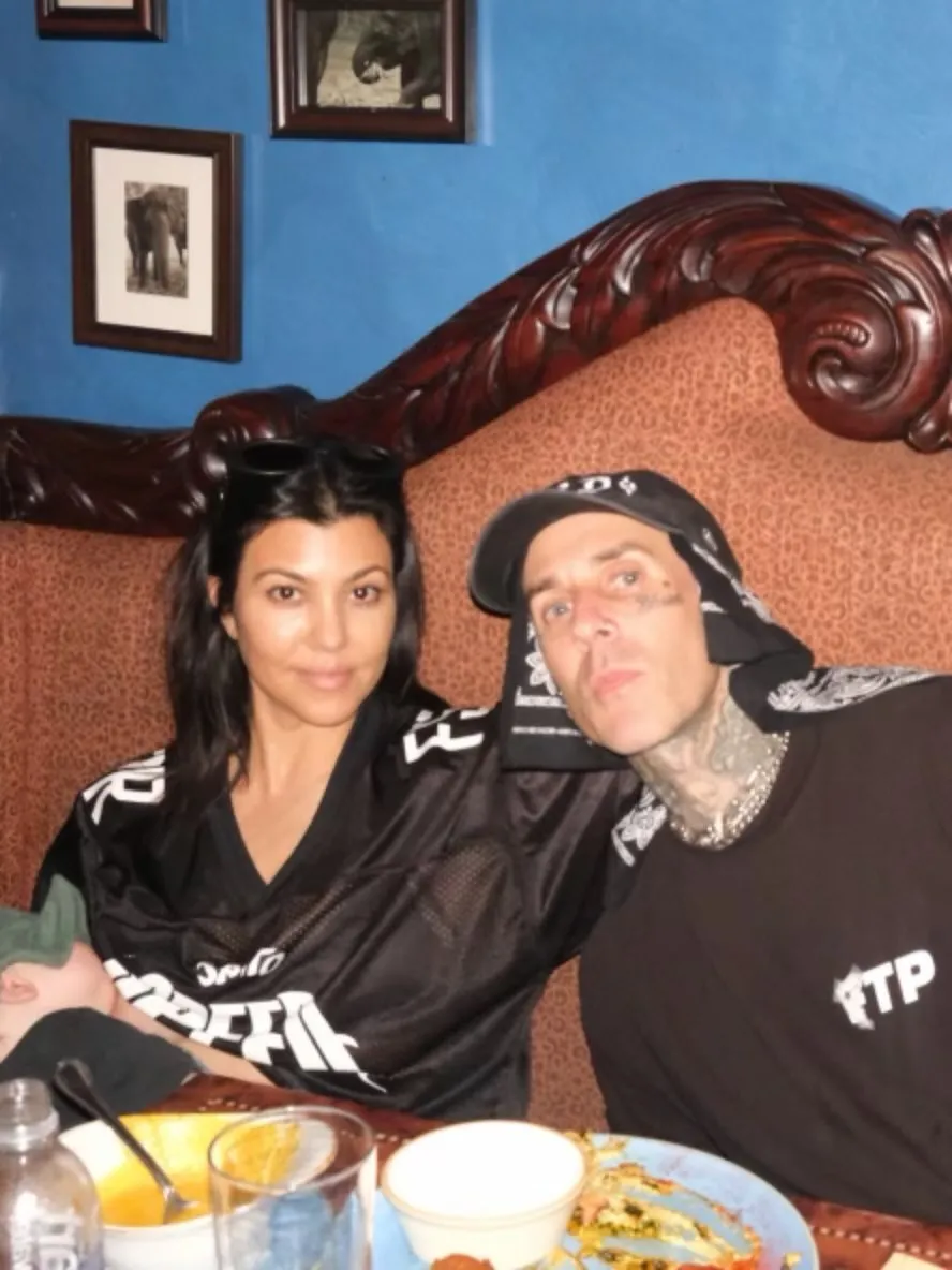 Kourtney Kardashian and Travis Barker sit at a table, with Barker's arm around Kardashian.