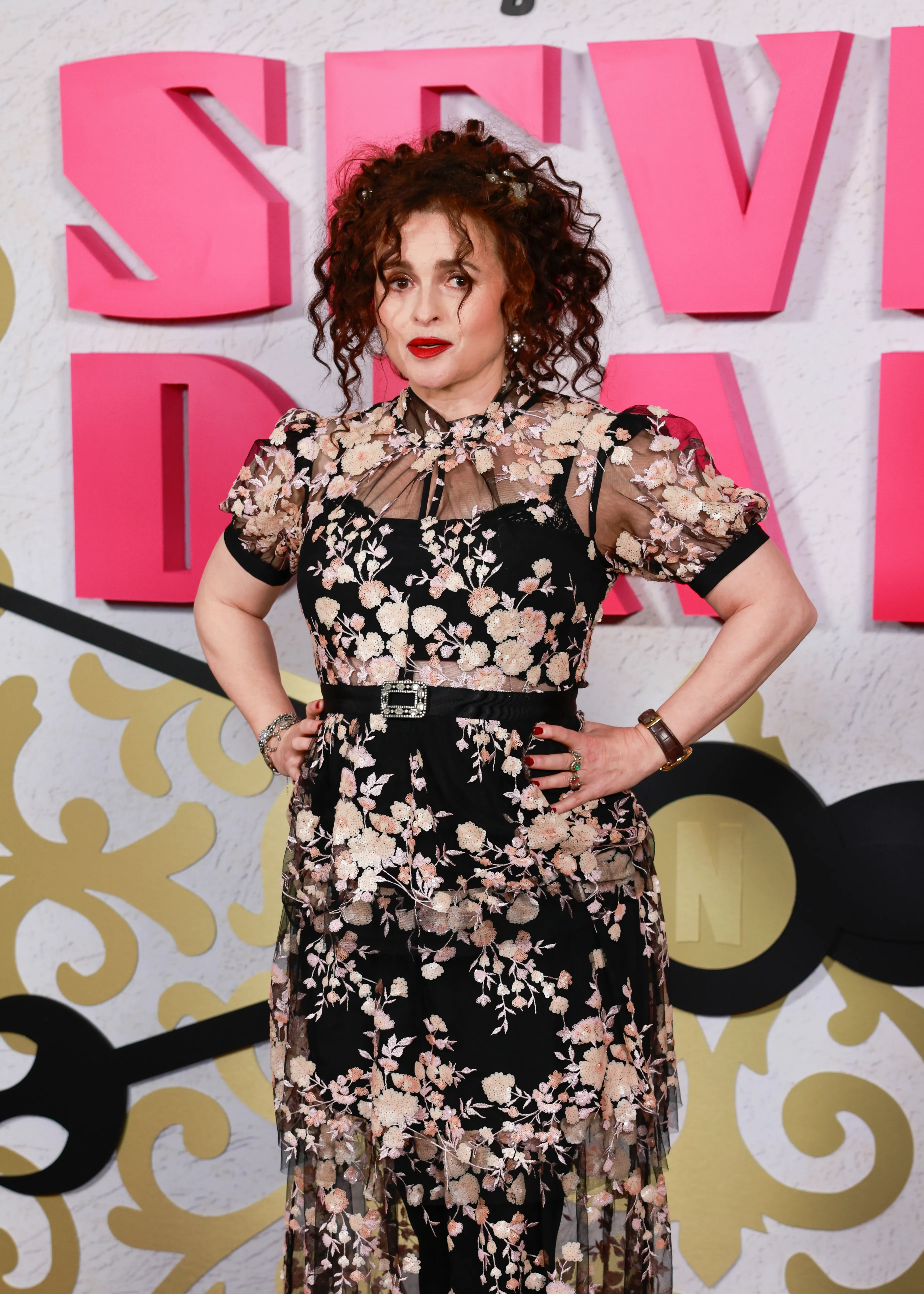 Helena Bonham Carter in a black embroidered sheer jacket and layered tulle skirt, with a colorful necklace and hair adorned with black and red accents, attending an event.
