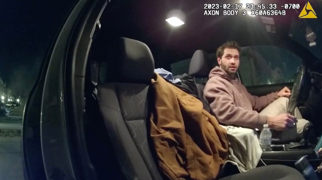 A man in a hoodie in a car, looking shocked.