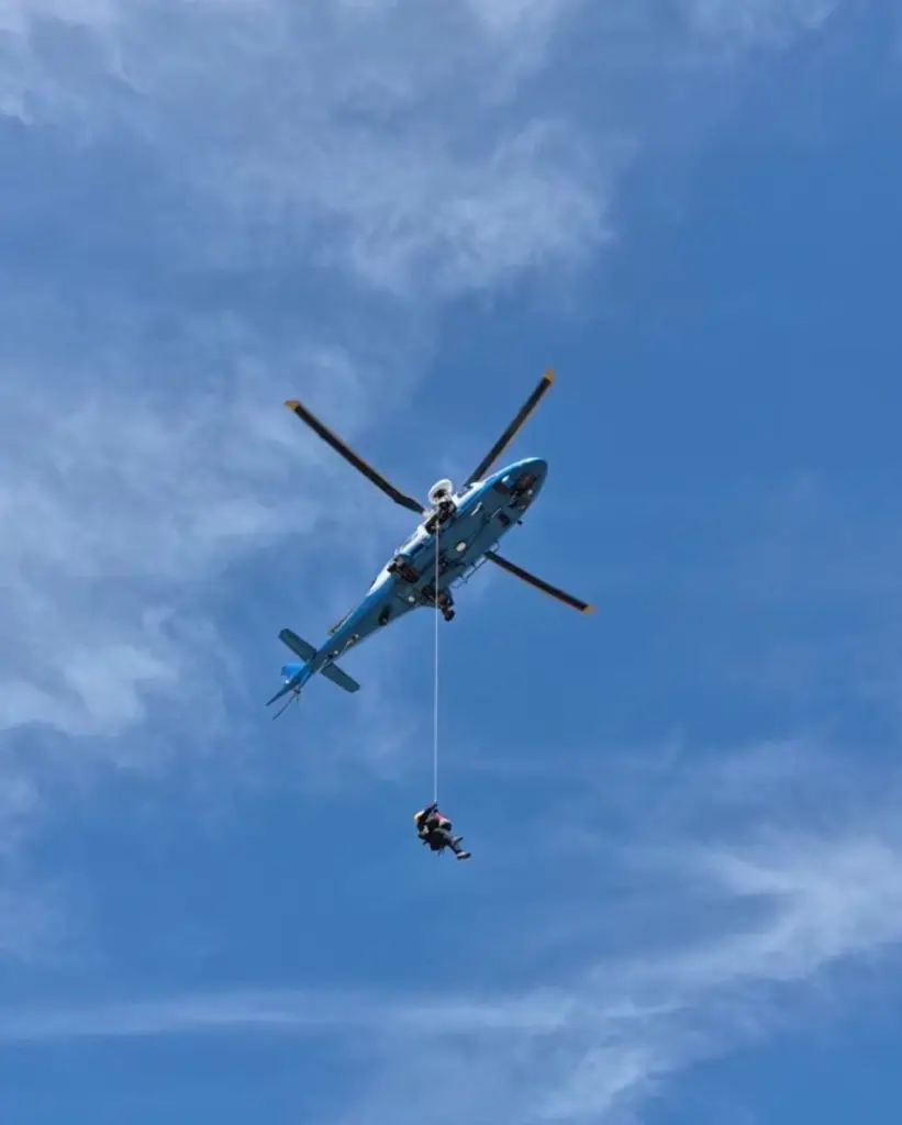 A blue helicopter hovers in the sky, lowering a person on a rope.
