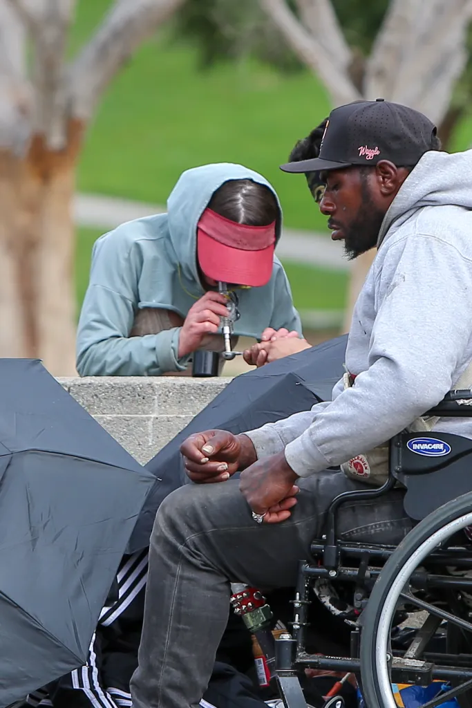Two unhoused people in MacArthur Park, one in a wheelchair, with umbrellas open.