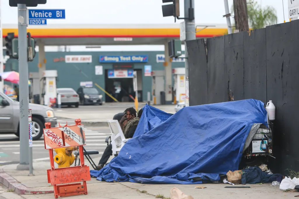 A person sits beside a blue tarp and shopping cart on the sidewalk of Venice Boulevard, with a gas station in the background.