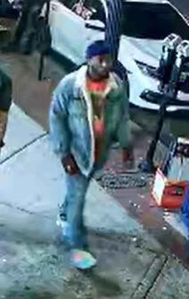 Still image of a male suspect wearing a blue denim jacket over an orange shirt, blue jeans, and a blue cap, walking on a sidewalk next to a white car.