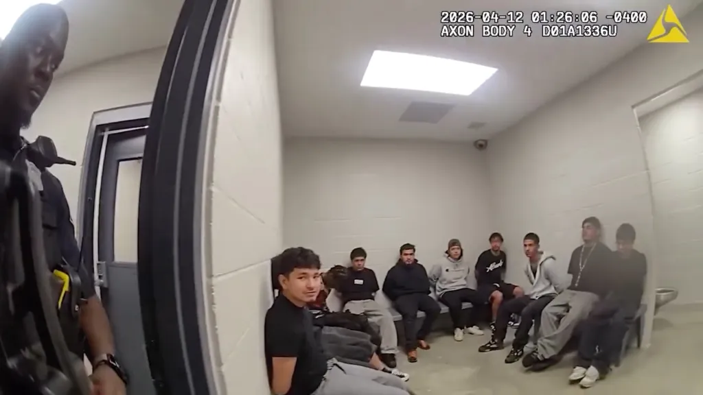 A police officer stands near an open door, while six men sit on a bench inside a detention room, with the time and date from a body camera visible in the top right.