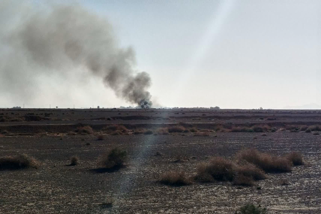 Black smoke rising from a distant site in a barren landscape under a bright sky, indicating an explosion or fire.