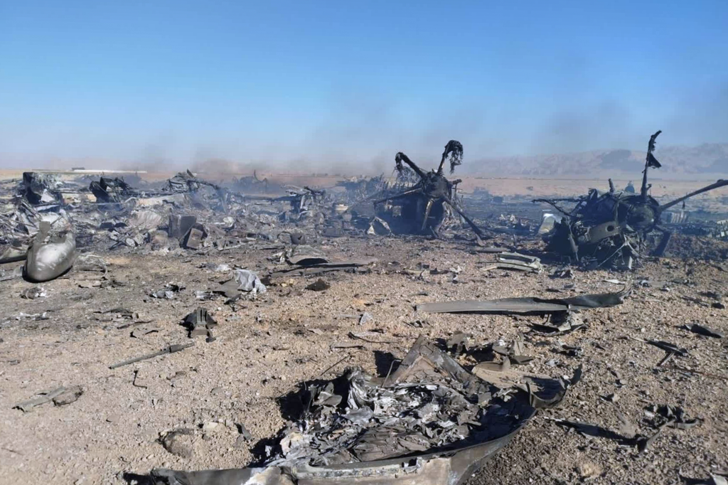 Wreckage of a downed American transport plane and two helicopters scattered across a desert landscape under a clear blue sky.