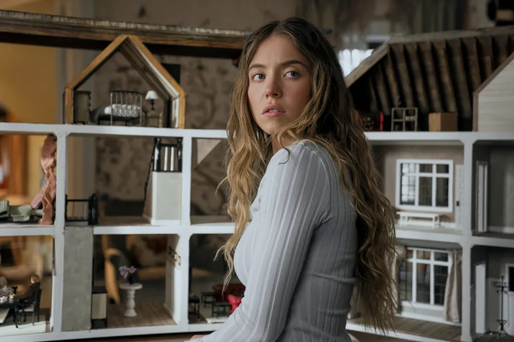 Sydney Sweeney in a light ribbed long-sleeved shirt, sitting in front of a large dollhouse, looking intently at the camera.