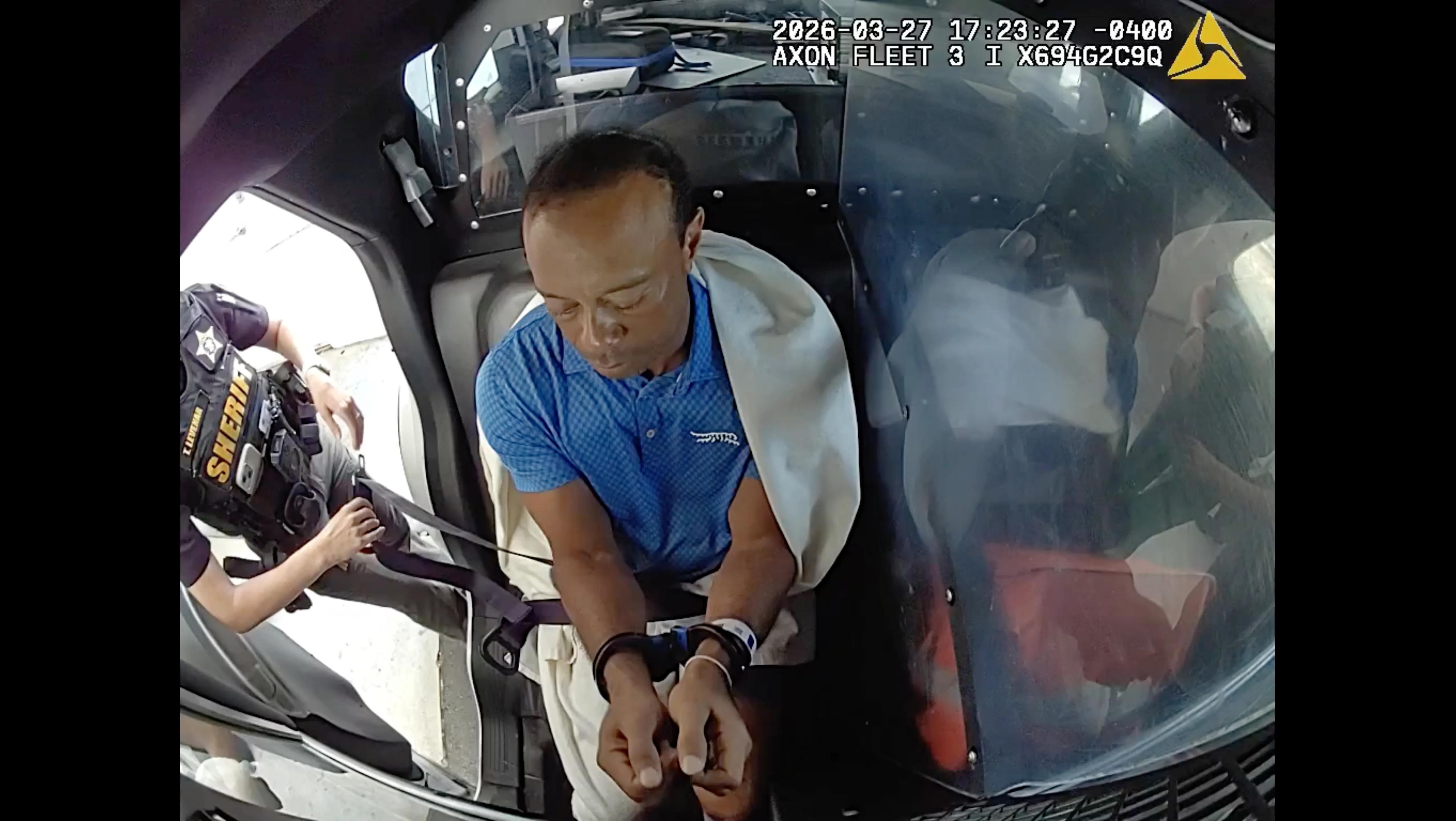 Tiger Woods in handcuffs, seated in the back of a police vehicle, with a white blanket over his shoulders.