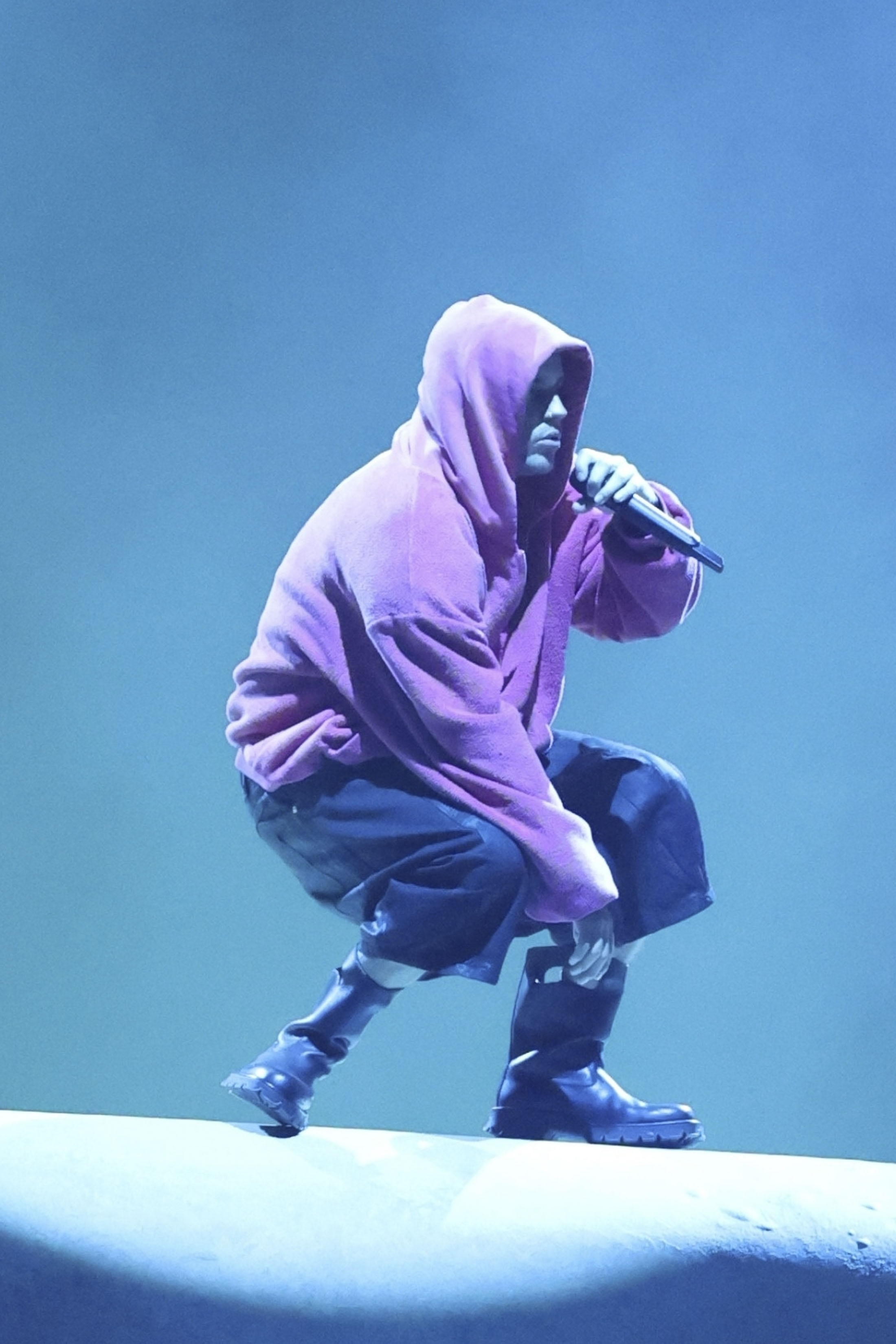 Justin Bieber performing at Coachella Music Festival, wearing a purple hoodie, black shorts, and black boots, holding a microphone.