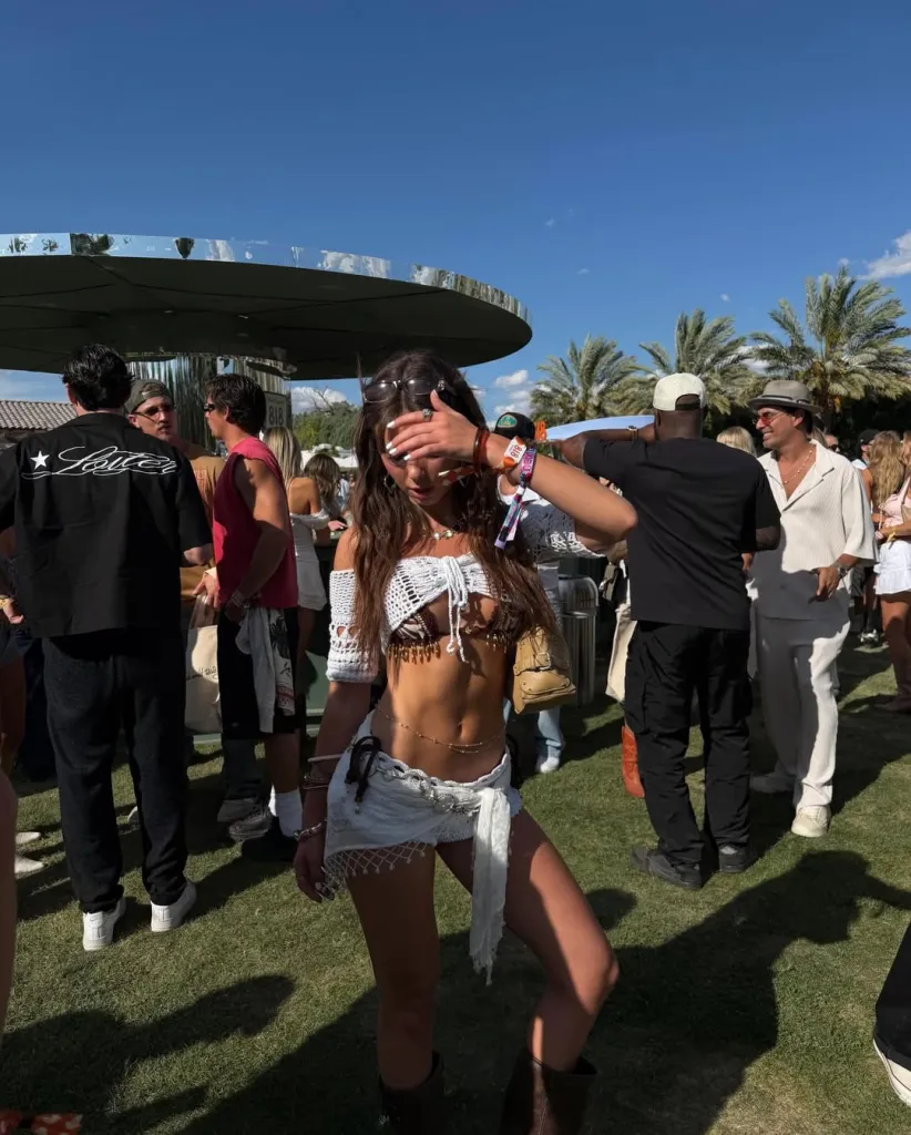 A woman in a crocheted top and white shorts shields her eyes from the sun in a crowded outdoor setting.