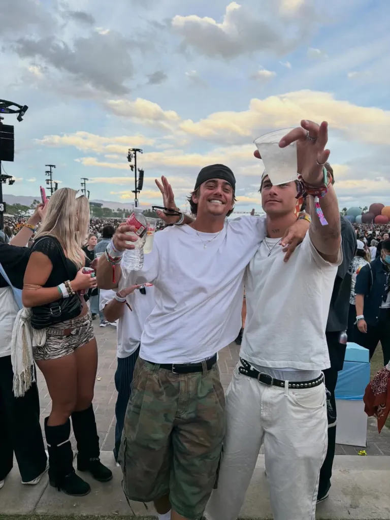 Two young men wearing white t-shirts and holding drinks, smiling in a crowd at an outdoor event.