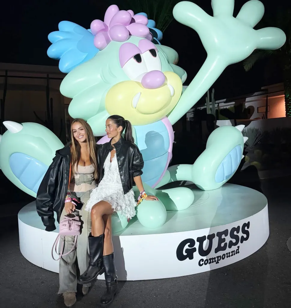 Two women in black jackets pose in front of a colorful, cartoonish Guess Compound mascot.