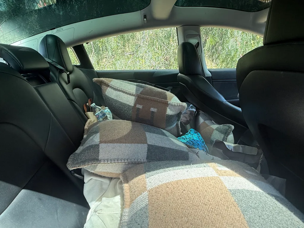 Backseat of an abandoned car with blankets, pillows, and water bottles, belonging to Sarinasadat Hosseiny.
