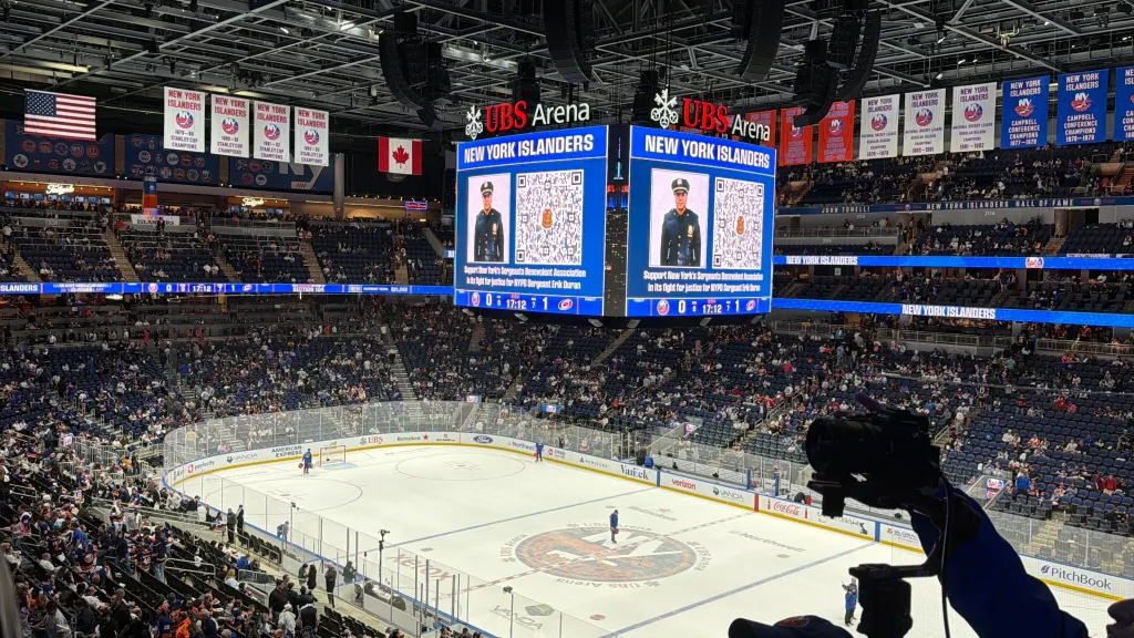An overhead view of a hockey game with two jumbotron screens showing a photo of NYPD Sergeant Erik Duran and a QR code for donations.