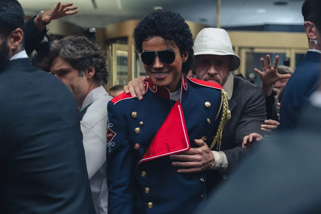 Jaafar Jackson as Michael Jackson in a red and blue military-style jacket, being escorted by KeiLyn Durrel Jones as Bill Bray.