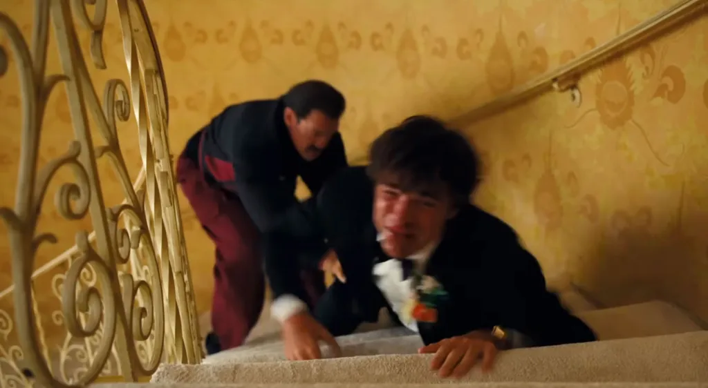 Man in tuxedo crying on stairs, with another man in a dark shirt and red pants behind him.