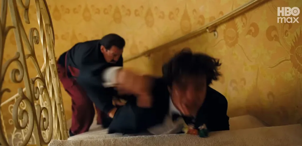 A man in a black suit with a mustache dragging a man in a tuxedo with messy hair up a carpeted staircase with a golden railing.