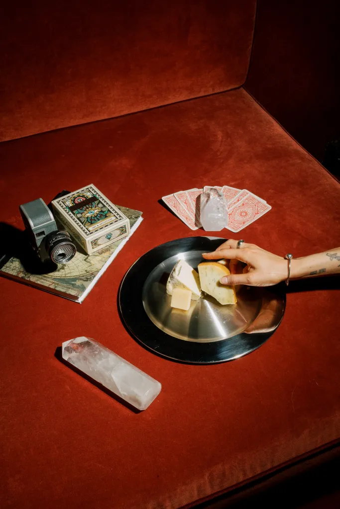 A hand selects a piece of cheese from a silver platter with other cheeses, next to crystals, playing cards, and a camera, all on a red velvet surface.