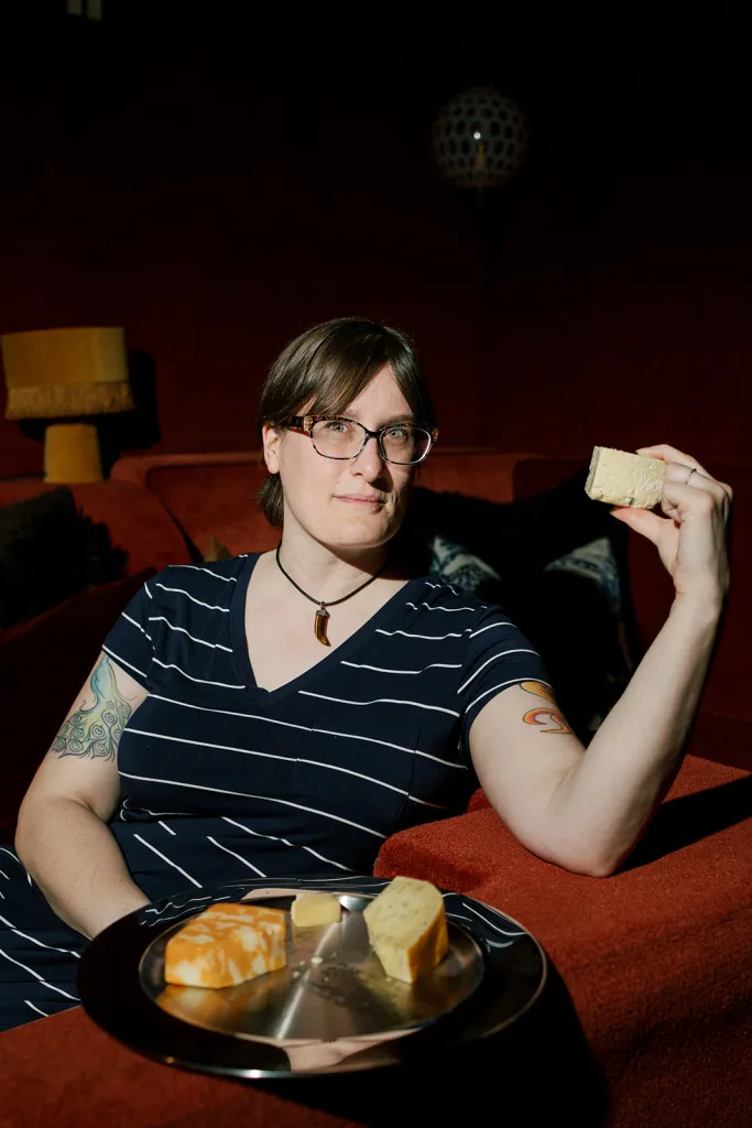Jennifer Billock, known as the Cheese Witch or Jen the Tyromancer, holds a piece of cheese while sitting on a red couch with a platter of cheese beside her.