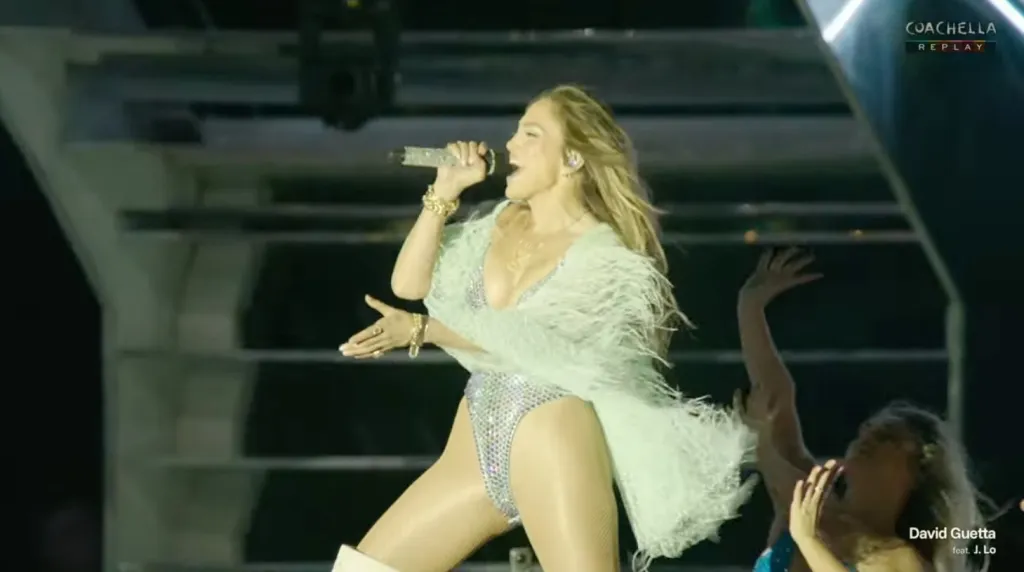 Jennifer Lopez performing on stage in a shimmering bodysuit with a feather boa, holding a microphone.