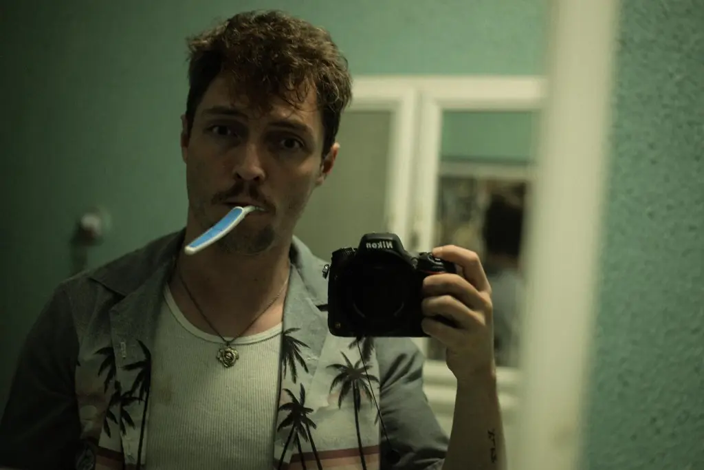 A man with a toothbrush in his mouth takes a mirror selfie while holding a camera.