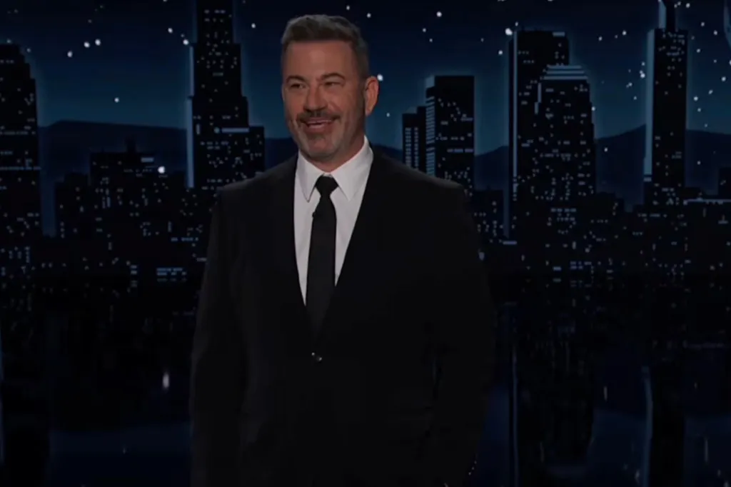Jimmy Kimmel smiling on stage in a black suit with a cityscape background.