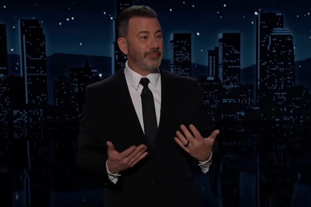 Jimmy Kimmel speaking on stage, wearing a black suit and tie, with a city skyline backdrop at night.