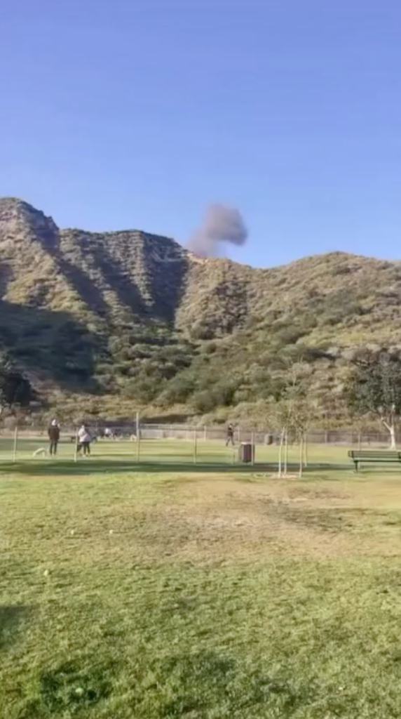 Smoke plume rising above a distant hillside, seen from a park.