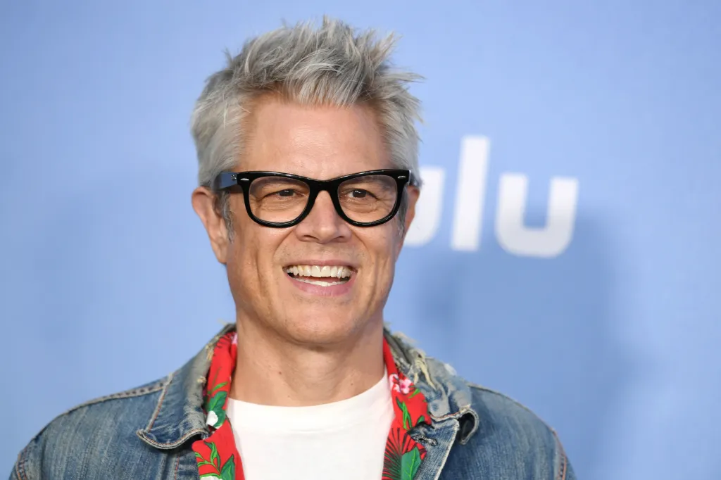 Johnny Knoxville in black glasses, a white t-shirt, and a denim jacket with a red shirt underneath, smiling against a blue background with the white 