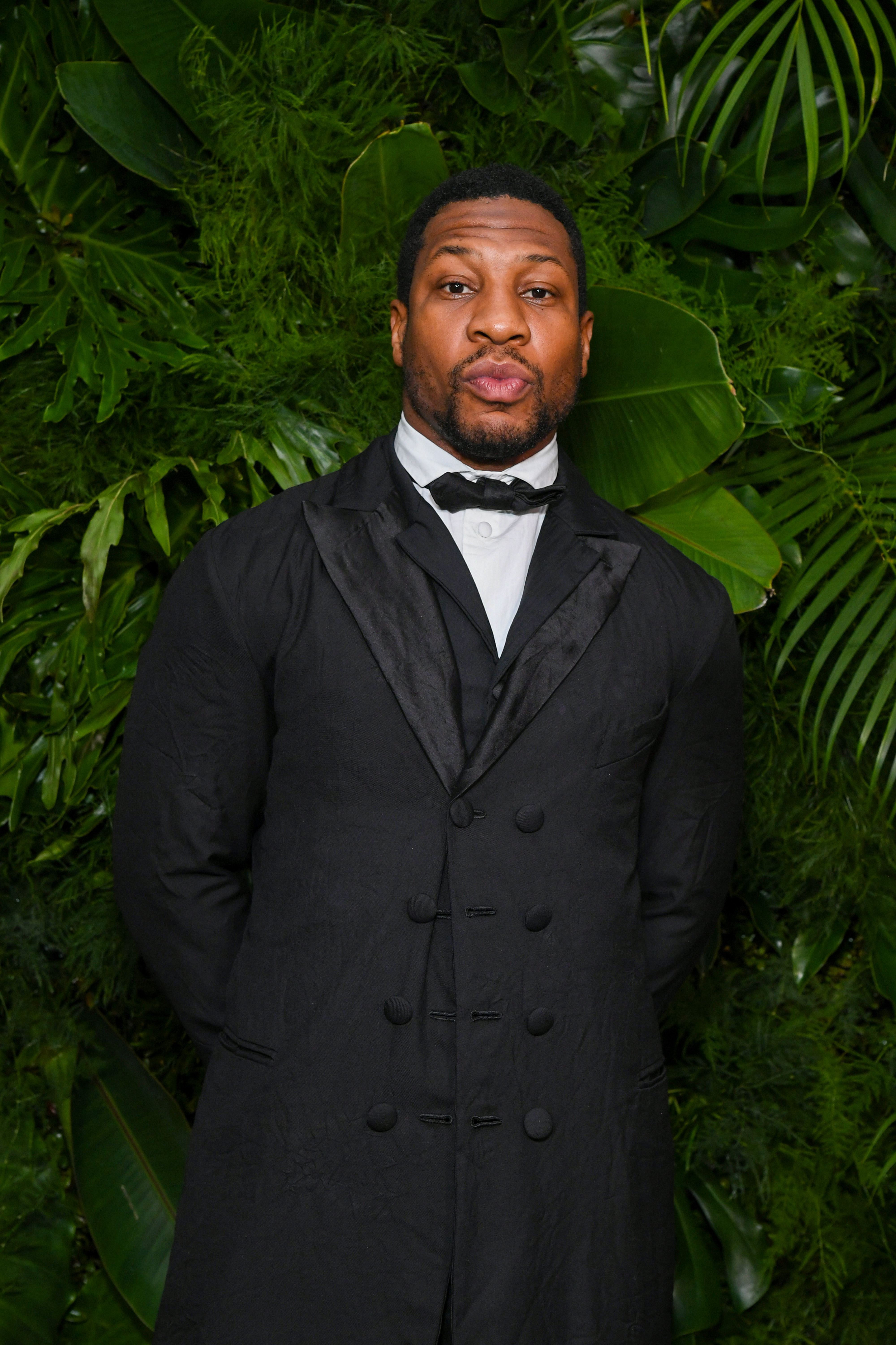 Jonathan Majors in a tuxedo against a background of green foliage.