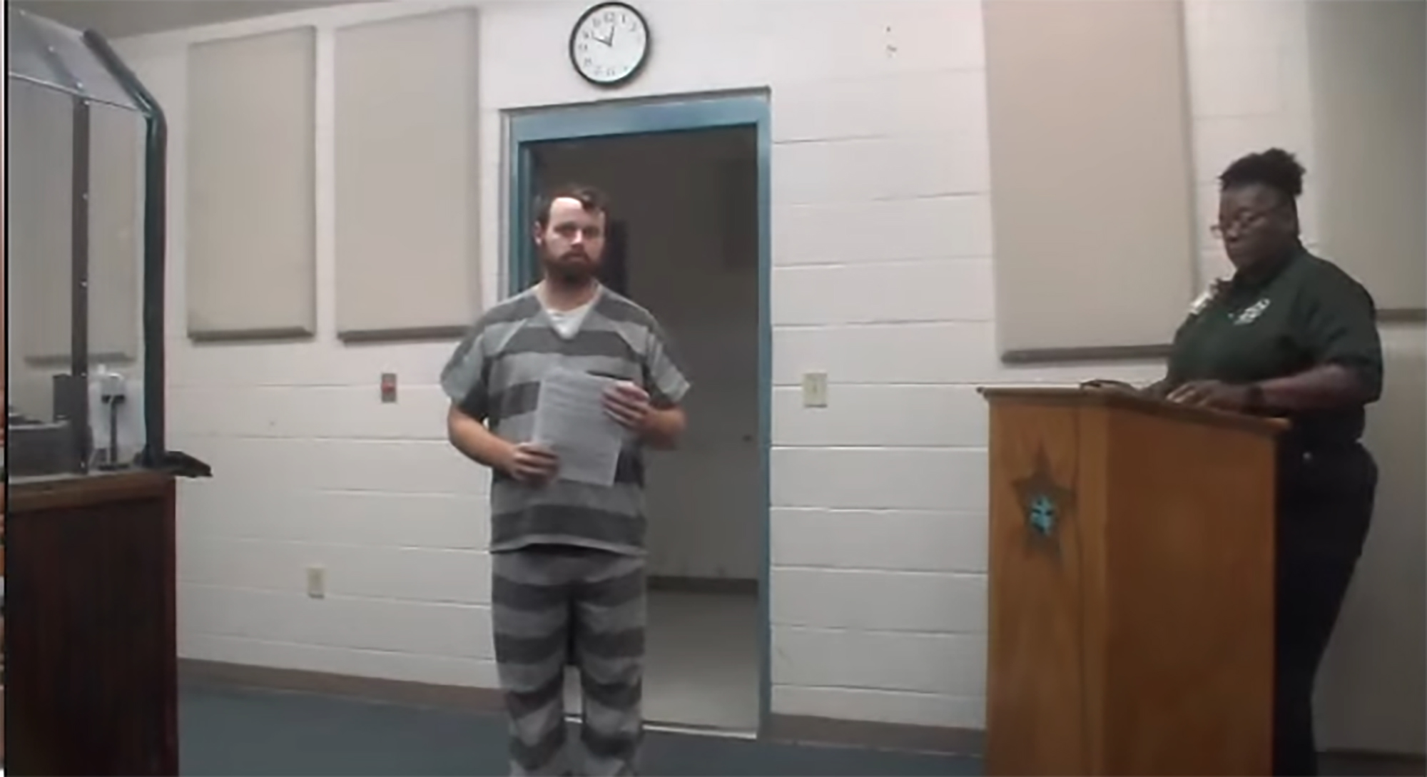 Joseph Duggar in court, wearing a striped prison uniform, holding papers, with an officer next to a podium.