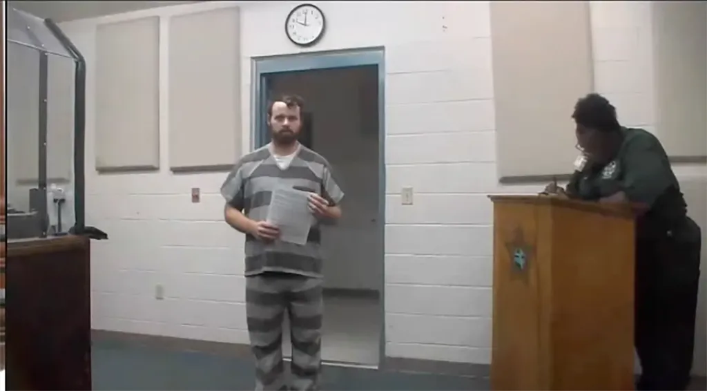 Joseph Duggar in a gray striped jail uniform holds papers during his first appearance at Bay County Jail.