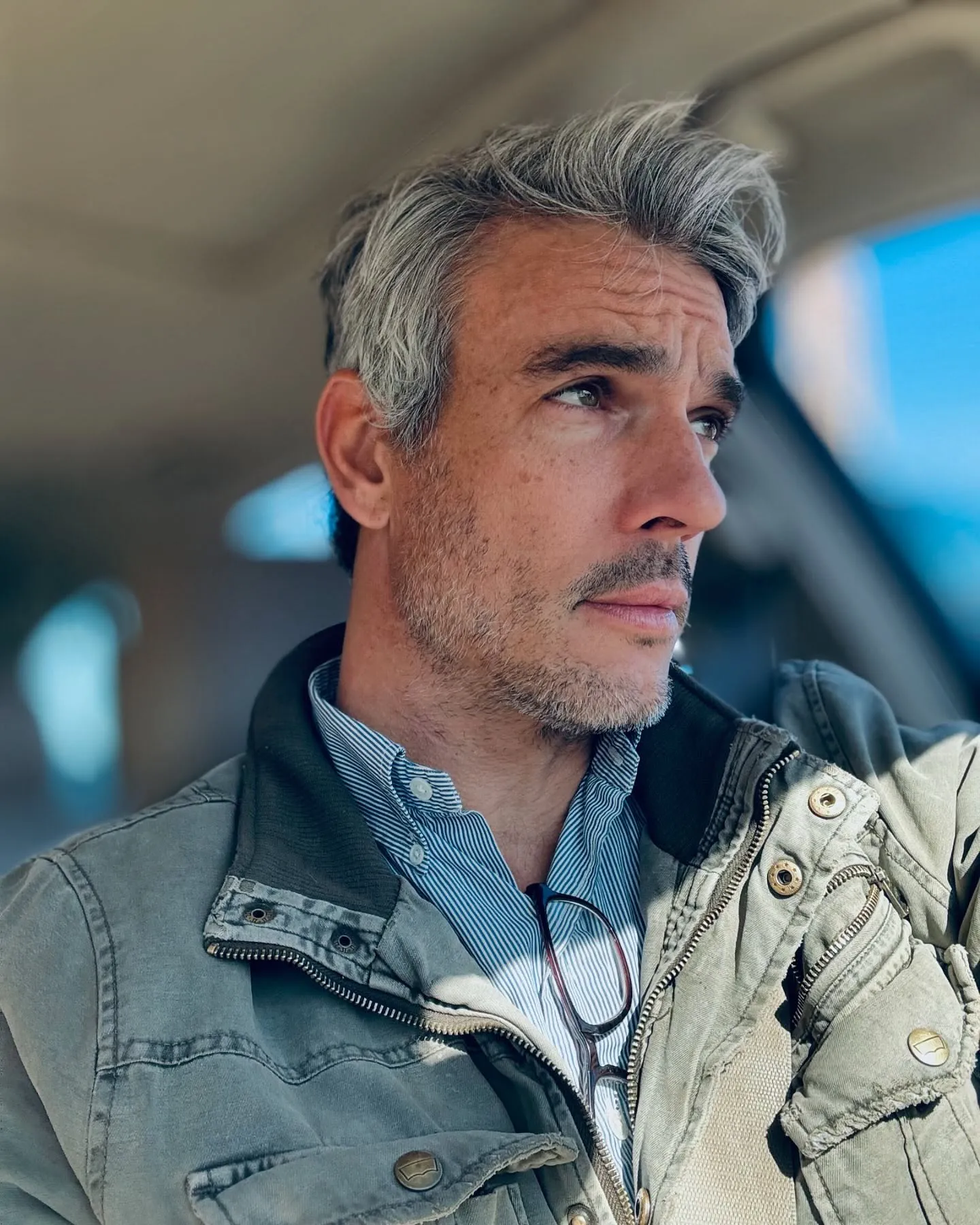 Josh Kloss with gray hair and a beard, wearing a striped shirt and green jacket, looking out a car window.