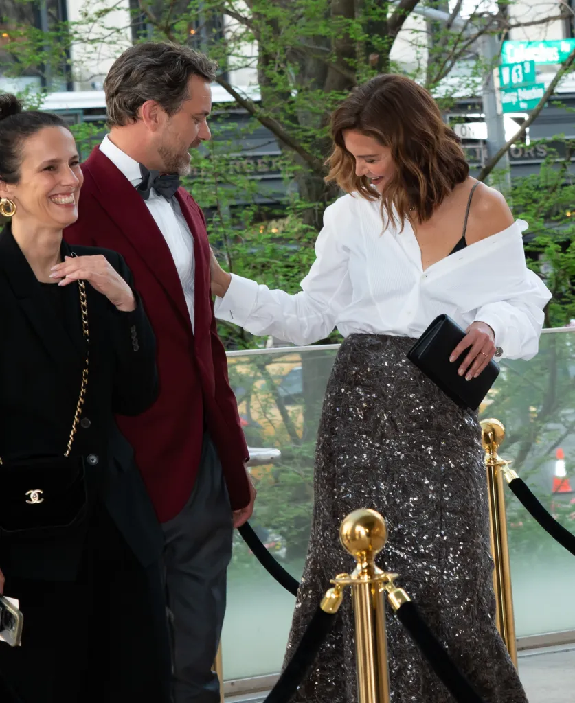 Katie Holmes in a white shirt and sparkling skirt, and Joshua Jackson in a burgundy tuxedo jacket, are seen on a red carpet with another woman.