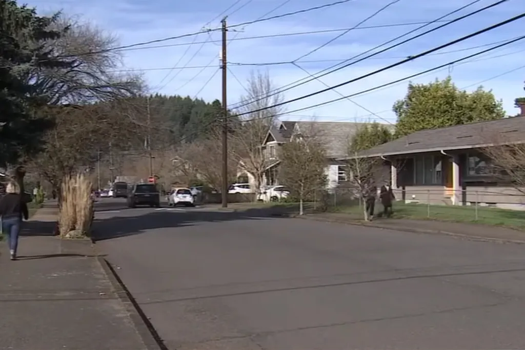 A woman walking on a sidewalk and two people walking on a street with houses and parked cars, and trees in the background.