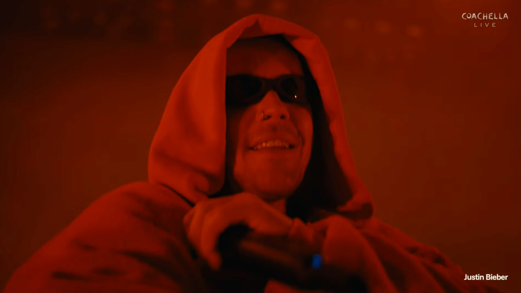 Justin Bieber on stage wearing a red hoodie and sunglasses, holding a microphone.