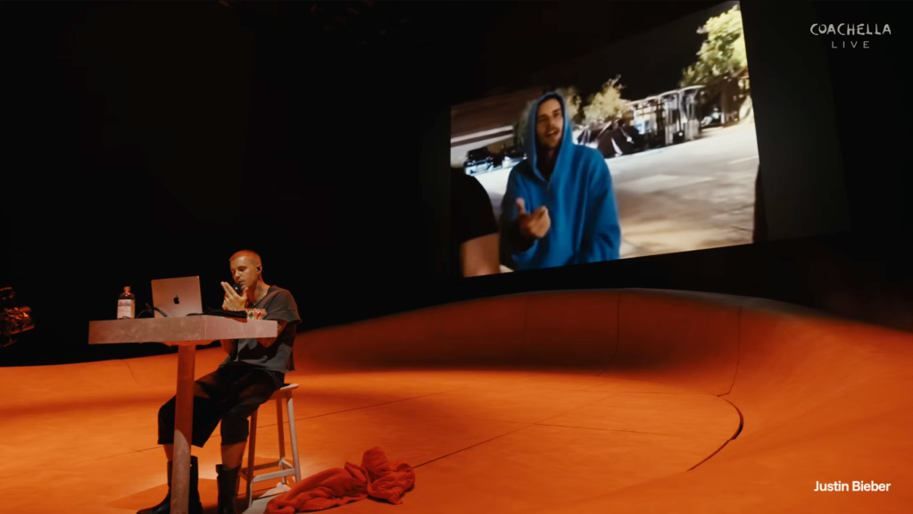 Justin Bieber performing on stage with a laptop, a large screen behind him displaying another person.