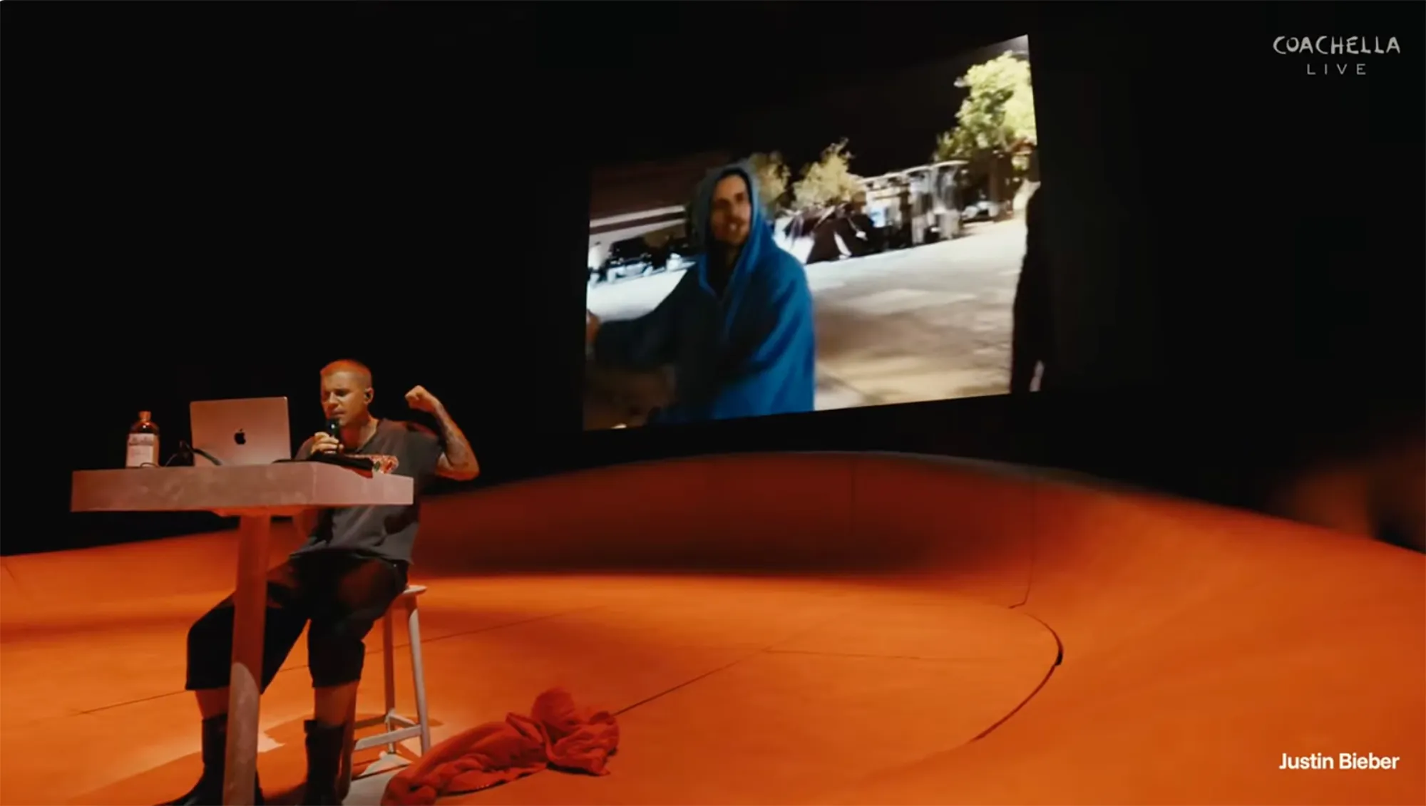 Justin Bieber sitting at a table with a laptop, microphone, and a bottle, performing on a stage with a screen showing a man in a blue hoodie.