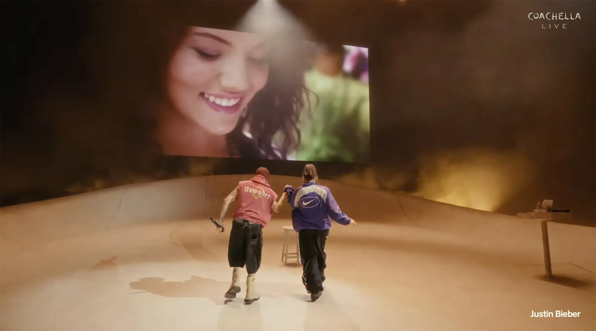 Justin Bieber and Billie Eilish on stage, holding hands, with a large video screen behind them showing a woman smiling.