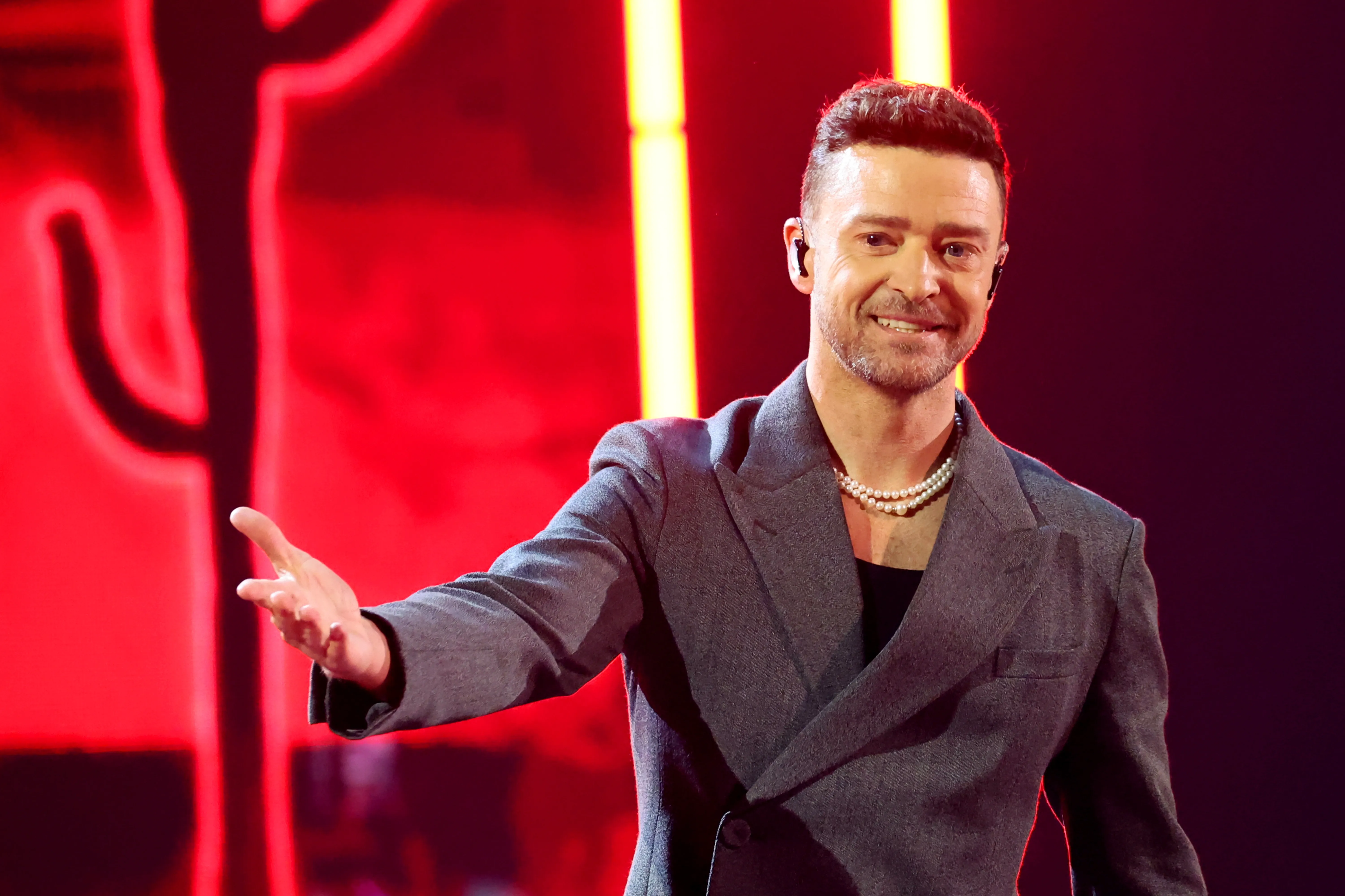 Justin Timberlake performing onstage, smiling at the audience with an outstretched arm, wearing a gray blazer and a pearl necklace.
