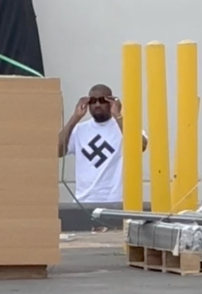 Kanye West wearing a white T-shirt with a swastika while adjusting his sunglasses.
