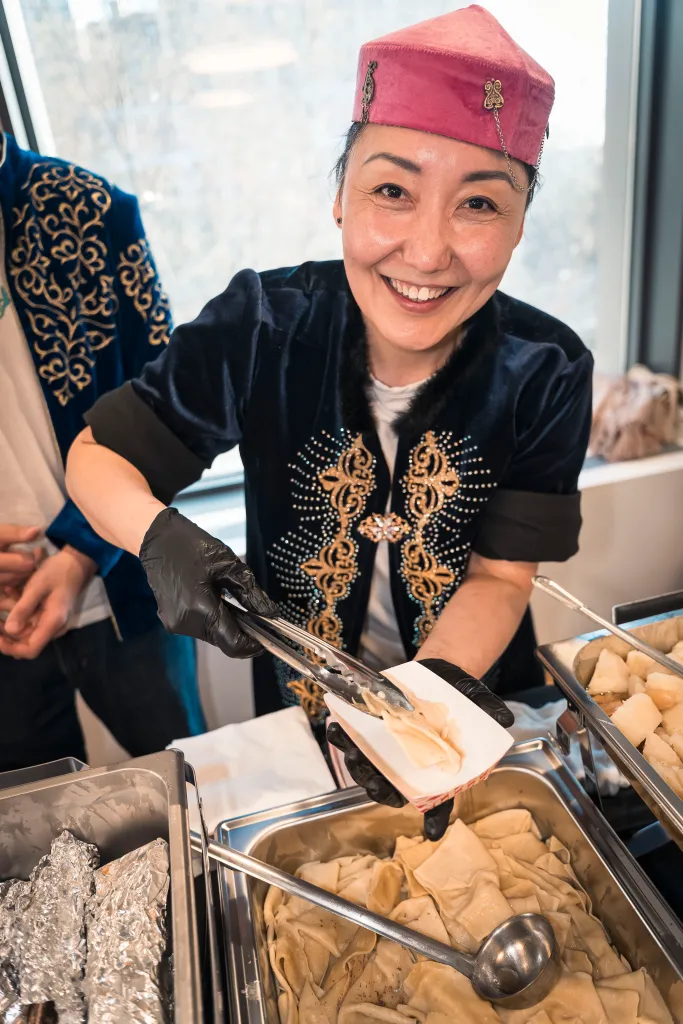 A smiling woman in traditional Kazakhstani clothing serving food from a buffet.