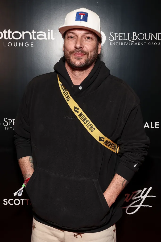 Kevin Federline posing in a black hoodie, beige shorts, and a white baseball cap.