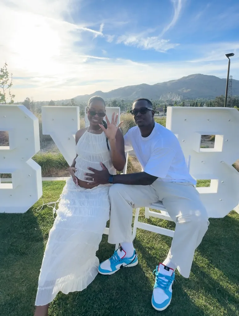 A pregnant woman and a man in matching white outfits sit together outdoors, in front of large white letters that spell 