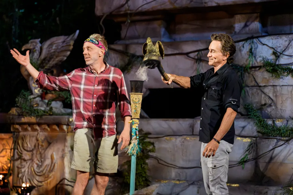 Two men, one holding a torch and the other gesturing, stand in front of an ancient-looking ruin.