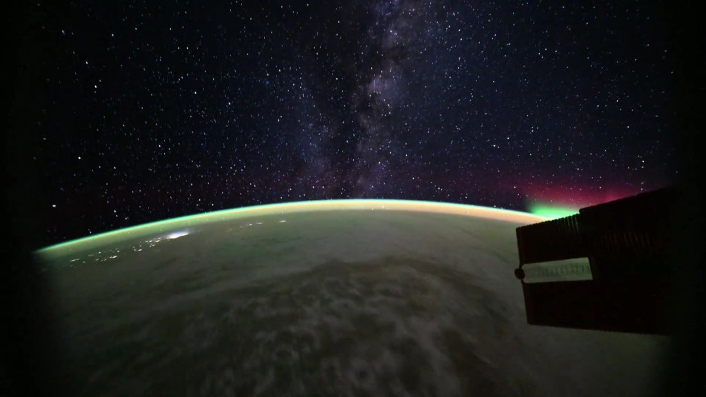 An aurora and the Milky Way seen from orbit, with Earth's curvature, city lights, and an orbital module in the foreground.